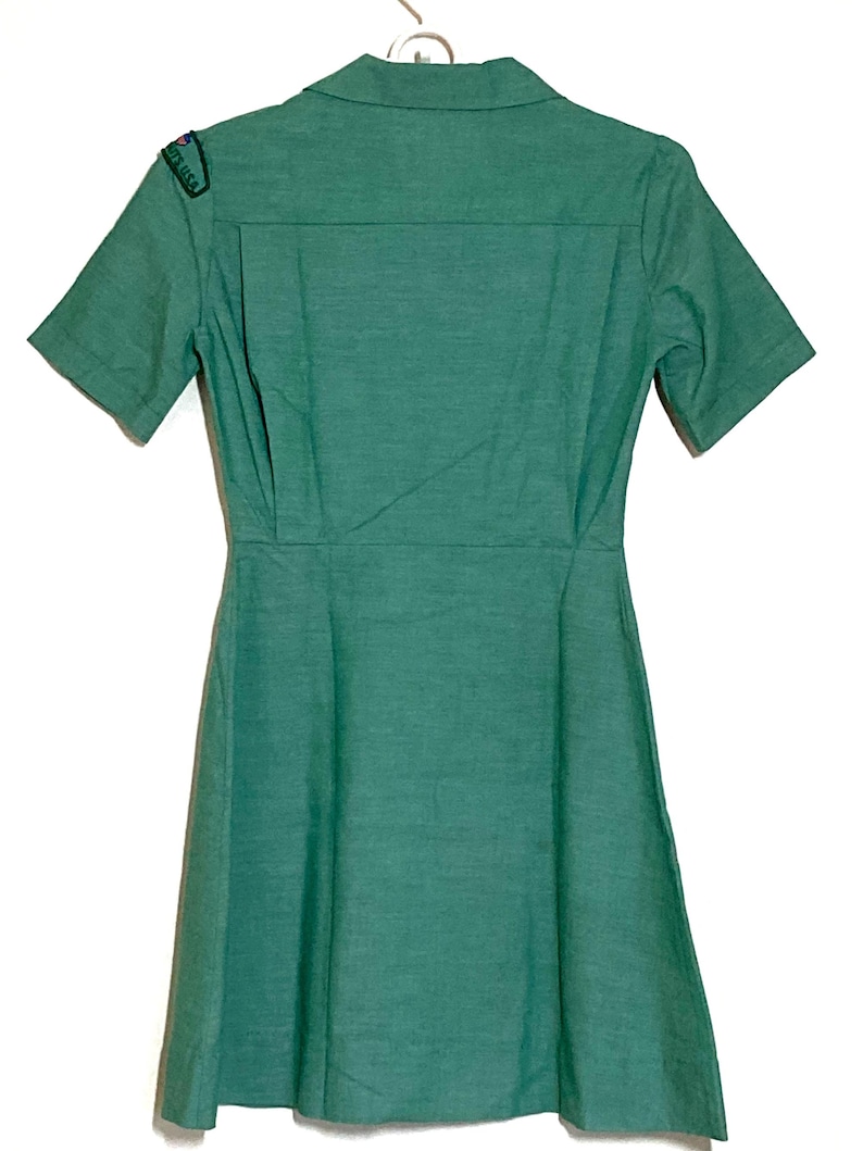 Puede incluir: Un vestido verde de manga corta con cuello y un parche en el hombro izquierdo que dice "Girl Scouts USA".