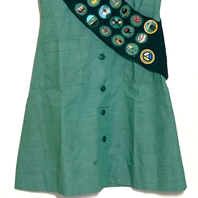 Puede incluir: Un vestido verde con una banda que presenta una variedad de insignias coloridas. El vestido tiene un frente abotonado y dos bolsillos.
