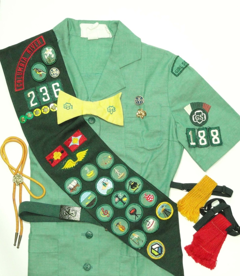 Puede incluir: Un uniforme verde de Girl Scout con una banda que presenta varias insignias y parches. La banda tiene el texto "Columbia River" y el n&uacute;mero "236". El uniforme tambi&eacute;n tiene una corbata de lazo amarilla y un pin de Girl Scout con el n&uacute;mero "188".