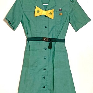 Puede incluir: Un vestido verde de uniforme de Girl Scout con mangas cortas, una corbata de lazo amarilla y un cintur&oacute;n. El vestido tiene un frente abotonado y dos bolsillos. El vestido tiene un parche en la manga derecha que dice "Girl Scout".