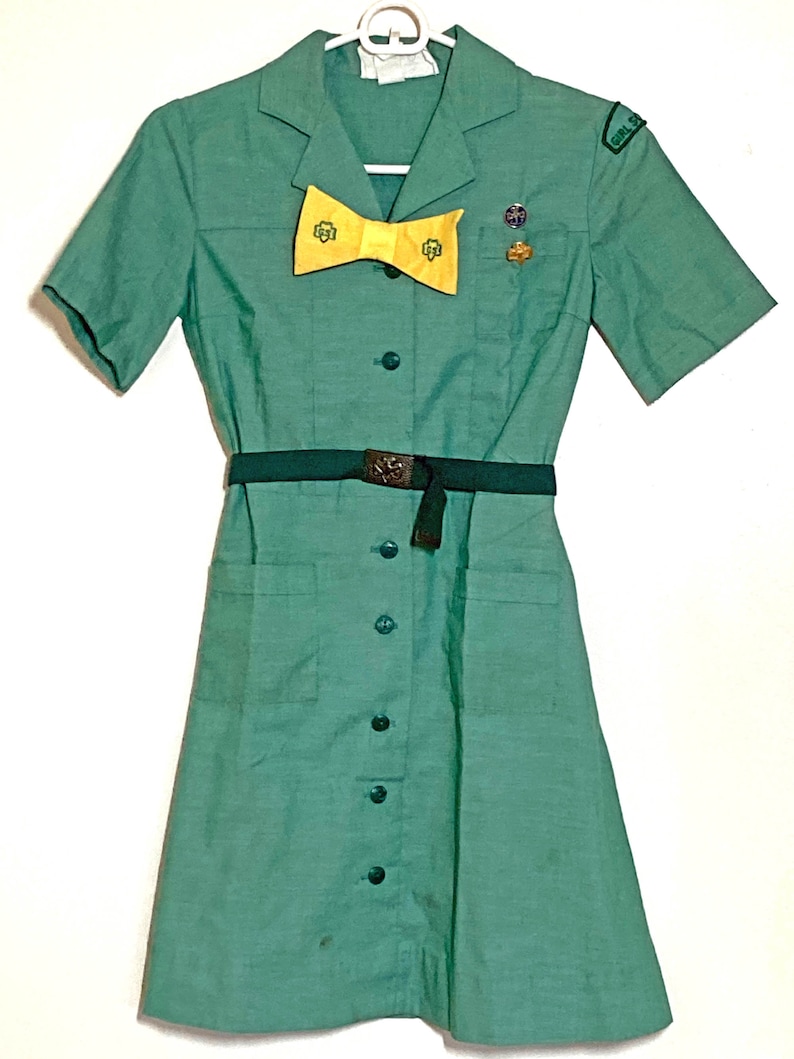 Puede incluir: Un vestido verde de uniforme de Girl Scout con mangas cortas, una corbata de lazo amarilla y un cintur&oacute;n. El vestido tiene un frente abotonado y dos bolsillos. El vestido tiene un parche en la manga derecha que dice "Girl Scout".