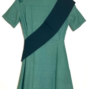 Puede incluir: Un vestido verde con mangas cortas y una faja negra. El vestido tiene cuello y cierre de botones.