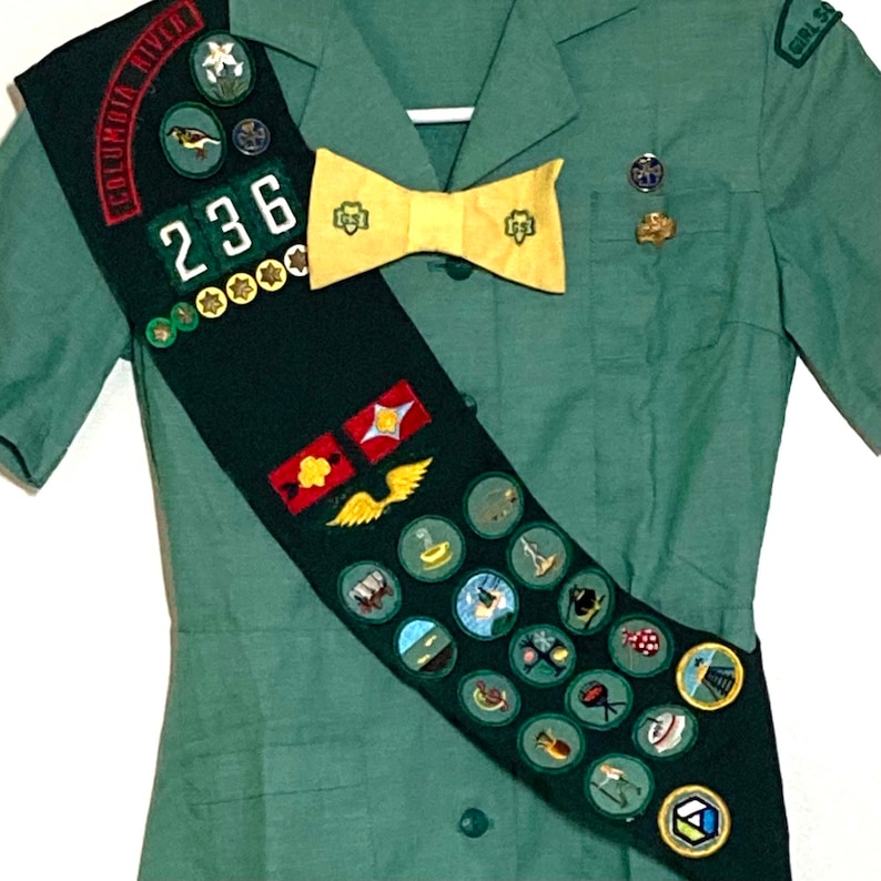 Puede incluir: Un uniforme verde de Girl Scout con una banda que presenta una variedad de insignias y una corbata de lazo amarilla. La banda tiene el texto "Columbia River" y el n&uacute;mero "236" en ella.