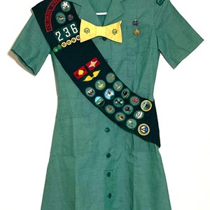 Puede incluir: Un uniforme verde de Girl Scout con una banda que presenta una variedad de insignias y una corbata de lazo amarilla. La banda tiene el texto "Columbia River" y el n&uacute;mero "236".