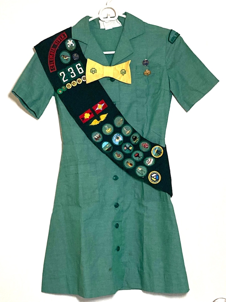 Puede incluir: Un uniforme verde de Girl Scout con una banda que presenta una variedad de insignias y una corbata de lazo amarilla. La banda tiene el texto "Columbia River" y el n&uacute;mero "236".