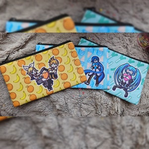 Peut inclure: Trois pochettes à fermeture éclair avec des motifs de personnages d'anime colorés. La première pochette a un fond jaune avec des fruits orange et des bananes jaunes. La deuxième pochette a un fond bleu avec un motif blanc et rose. La troisième pochette a un fond vert avec un motif blanc et rose.