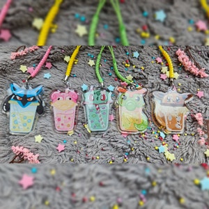 May include: Five colorful character charms in the shape of a boba tea cup. Each charm features a different Pokemon character, including Bulbasaur, Squirtle, Charmander, Pikachu, and a green dragon. The charms are attached to colorful strings.