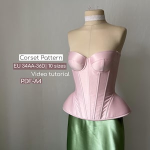 Op de afbeelding: Een lichtroze corset op een paspop, met een gestructureerd ontwerp met verticale naden en een peplum. De afbeelding bevat tekst: "Corset Pattern EU 34AA-36D | 10 sizes Video tutorial PDF-A4". De paspop draagt een groene satijnen rok.