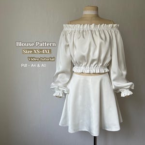 以下が含まれることがあります： マネキンに展示された、白いサテンのオフショルダーブラウスとマッチングスカートのセット。ブラウスは、フリル付きの袖口とフリル付きのネックラインが付いた長袖です。スカートはミニスカートです。画像には「Blouse Pattern Size XS-4XL Video tutorial Pdf - A4 & A0」と書かれています。