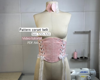 May include: A pink corset belt with white lacing and ribbon details is displayed on a mannequin. The belt is part of a pattern, with sizes ranging from XXS to XXL. The image includes text indicating a video tutorial and PDF A4 format.