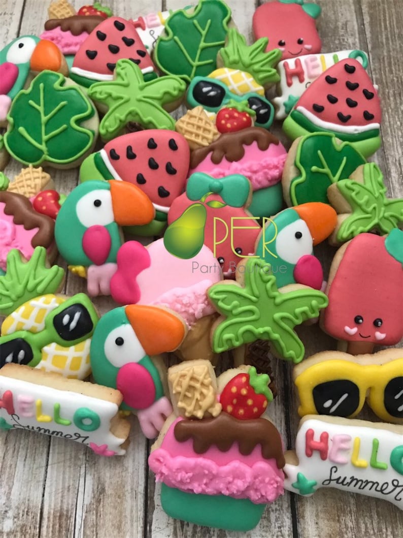May include: A collection of colorful, decorated sugar cookies in a summer theme. Cookies include toucans, watermelon slices, sunglasses, ice cream, and tropical leaves. Some cookies have the words "Hello Summer" written on them. The cookies are arranged on a light brown wooden surface.