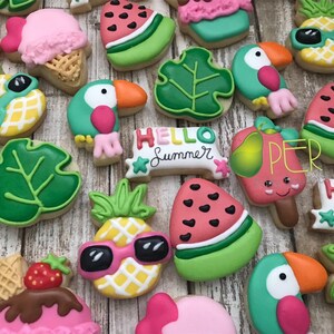 May include: A collection of decorated sugar cookies with a summer theme. The cookies are shaped like pineapples with sunglasses, toucans, watermelon slices, ice cream cones, and tropical leaves. Some cookies have the text "Hello Summer".