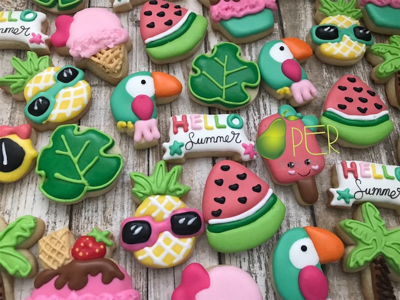 May include: A collection of decorated sugar cookies with a summer theme. The cookies are shaped like pineapples with sunglasses, toucans, watermelon slices, ice cream cones, and tropical leaves. Some cookies have the text "Hello Summer".