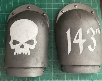May include: Two gray metal shoulder pads with white painted details. One pad has a skull design and the other has the number "143" painted on it.