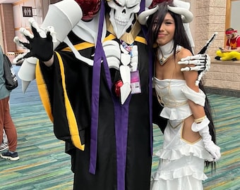 May include: Two people in cosplay costumes. One is a skeleton-like figure in a black robe with purple accents and a gold crown. The other is a woman in a white dress with long black hair and white gloves. They are posing for a photo.