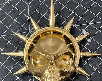 Op de afbeelding: Een gouden schedel met een halo van spikes. De schedel heeft een gedetailleerd gezicht met oogkassen, een neus en een mond. De halo is gemaakt van een cirkel met spikes die naar buiten stralen.