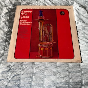 May include: A vintage vinyl record album cover featuring a red background with the title "Tubby The Tuba & Other Children's Favorites". The cover features a cartoon illustration of a tuba with a drum in front of it.