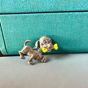 May include: A silver-toned dog brooch with yellow enamel bone accents. The dog is depicted running and holding the bone in its mouth.