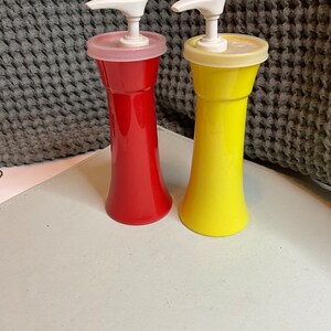 May include: Two plastic condiment dispensers with pumps. The dispenser on the left is red and the dispenser on the right is yellow.