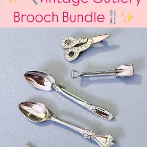 May include: A set of five vintage silver-toned cutlery brooches, including a pair of scissors, a shovel, and three spoons. The brooches are adorned with rhinestones and have intricate details.
