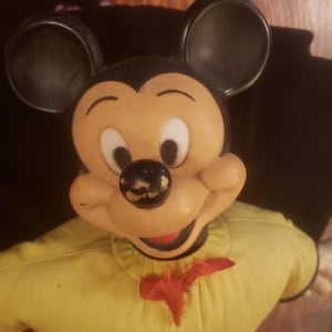 May include: A vintage Mickey Mouse plush toy with black ears, a yellow shirt, and red shorts. The toy has a black nose and a friendly expression.