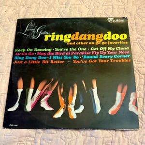 May include: Vinyl record album cover featuring a group of people wearing white boots with colorful accents. The album title is "Ring Dang Doo" and other au go go favorites by The Living Guitars. The album cover is black with colorful text and images.