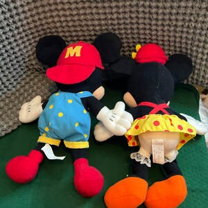 Peut inclure: Une paire de peluches Mickey Mouse et Minnie Mouse. Mickey porte une casquette rouge avec un "M" jaune et une salopette bleue &agrave; pois jaunes. Minnie porte un n&oelig;ud rouge et une jupe &agrave; pois jaunes et rouges.