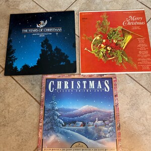 May include: Three vintage Christmas record albums. One album has a starry night sky design with text that reads "The Stars of Christmas". Another album has a red background with a horn and Christmas decorations. The third album has a winter scene with the word "Christmas" and "Listen to the Joy".