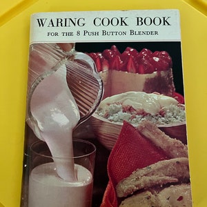 May include: A vintage Waring Cook Book for the 8 Push Button Blender. The cover features a photo of a glass of milk being poured, a bowl of salad, and a plate of fried chicken.