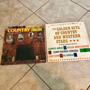 May include: Two vintage vinyl record albums. One album cover reads "COUNTRY Collection" with images of country singers. The other album cover reads "THE GOLDEN HITS OF COUNTRY AND WESTERN STARS" with a list of artists.