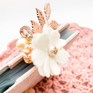 May include: A decorative floral embellishment featuring a cream-colored fabric flower with a pearl center, accented by gold-toned leaves and a cluster of beads. The embellishment is attached to a small, light pink book.