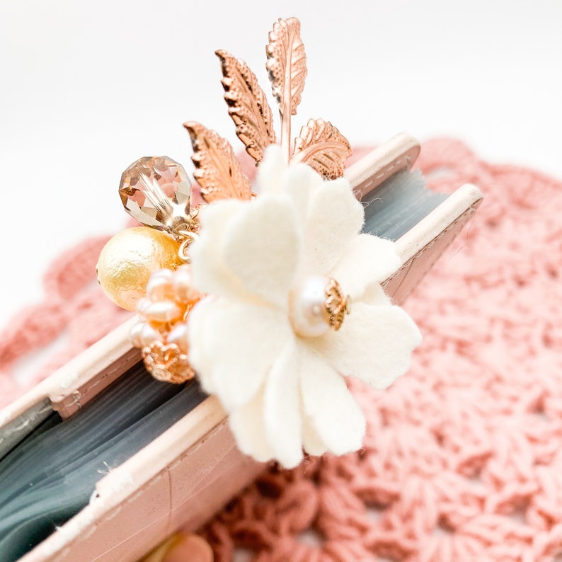 May include: A decorative floral embellishment featuring a cream-colored fabric flower with a pearl center, accented by gold-toned leaves and a cluster of beads. The embellishment is attached to a small, light pink book.