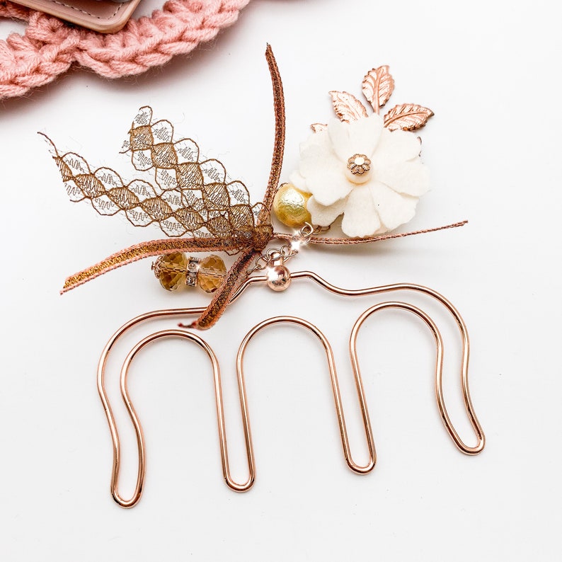 May include: A rose gold hair comb with a decorative floral and lace design. The comb features a white fabric flower, gold-colored beads, and a delicate lace accent. The hair accessory is designed for formal occasions.