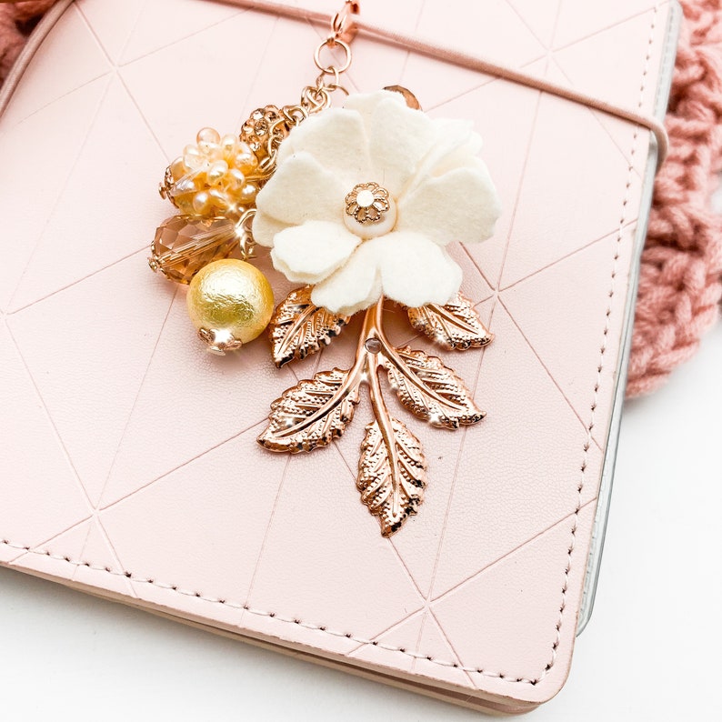 May include: A decorative charm featuring a cream-colored felt flower with a gold center, gold leaves, and various beads. The charm is attached to a pink leather notebook. The charm is a beautiful accessory for journals and planners.
