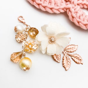 May include: A decorative charm with a rose gold clasp, featuring a white felt flower with a rhinestone center, rose gold leaves, and various gold and pearl beads. A pink knitted item is in the background.