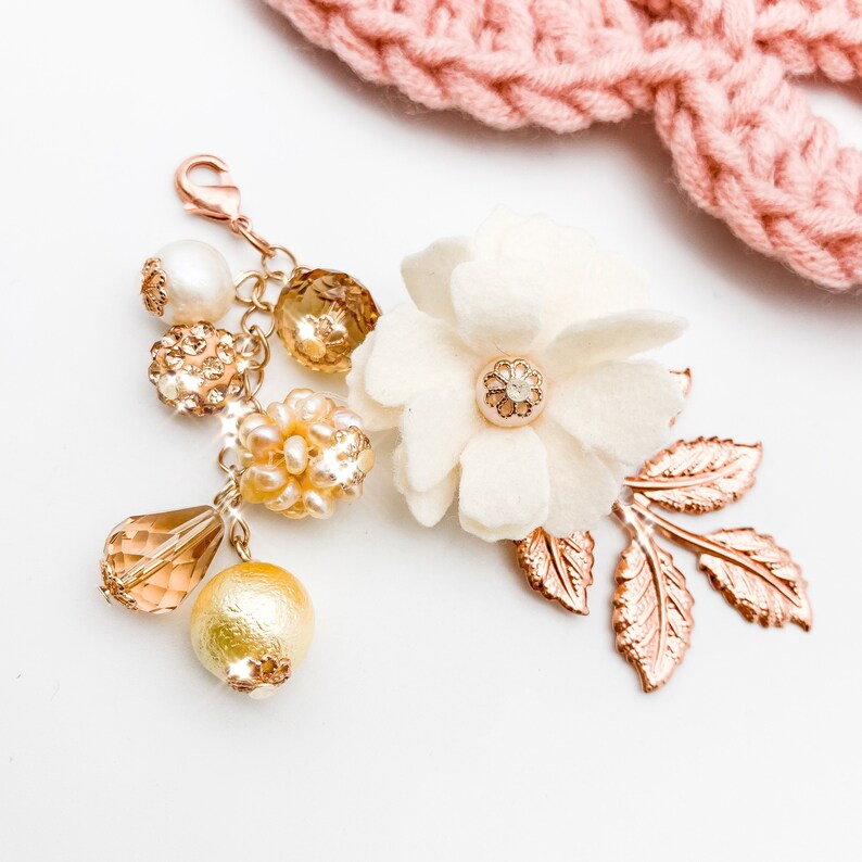 May include: A decorative charm with a rose gold clasp, featuring a white felt flower with a rhinestone center, rose gold leaves, and various gold and pearl beads. A pink knitted item is in the background.