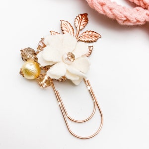 May include: A decorative paper clip featuring a cream-colored felt flower with a pearl center, surrounded by gold-toned beads and leaves. The clip is made of rose gold metal, adding a touch of elegance to stationery or office supplies.