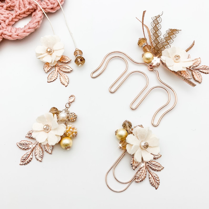 May include: A collection of floral hair accessories and jewelry. The pieces include cream-colored fabric flowers, gold-colored metal leaves, and various beads and pearls. The accessories are arranged on a white surface.