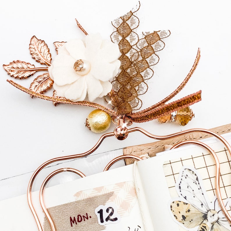 May include: A decorative rose gold metal clip with a cream-colored flower, gold bead, and brown lace and ribbon accents. The clip is attached to a notebook with a butterfly illustration and the date "MON. 12".