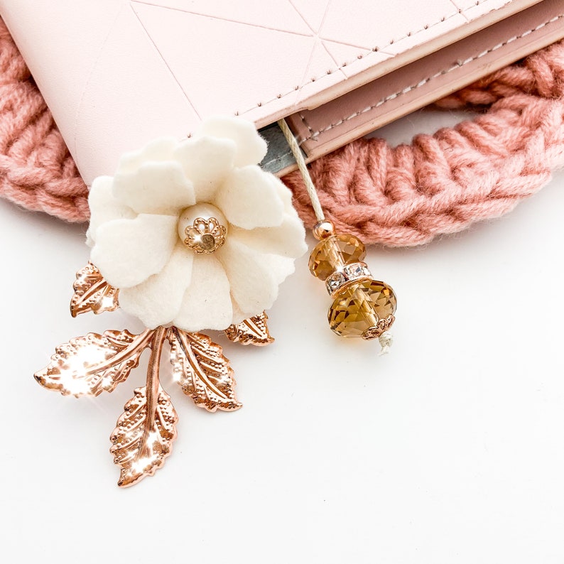 May include: A close-up of a handmade bookmark with a white felt flower, rose gold leaves, and a beaded tassel. The bookmark is next to a pink planner and a knitted pink headband. The flower has a gold center.