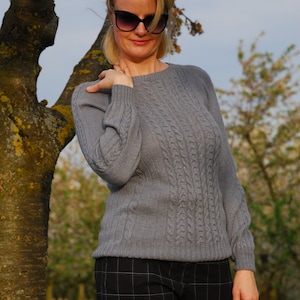 May include: A woman wearing a gray cable knit sweater and black and white plaid pants. She is standing in front of a tree with pink blossoms.