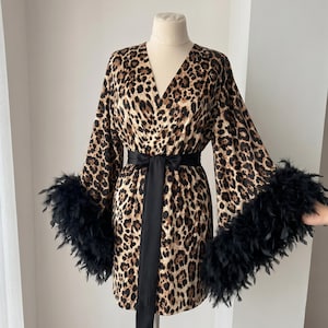 Puede incluir: Bata con estampado de leopardo con cinturón negro y ribete de plumas negras en las mangas. La bata se exhibe en un maniquí. La bata tiene cuello en V y mangas largas.