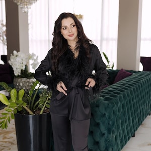 May include: A black satin pyjama set with feather trim. The trousers have a wide leg and the top has a shawl collar. The set is worn by a person standing in a room with a green sofa.