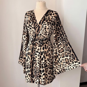 May include: A leopard print wrap dress with long, flared sleeves. The dress has a V-neck and a matching tie belt. The fabric features black spots on a tan background. The dress is displayed on a mannequin.