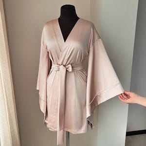 May include: A blush-colored satin robe with wide sleeves and a matching belt. The robe is displayed on a black mannequin, showcasing its flowing design and elegant drape. The fabric has a subtle sheen.