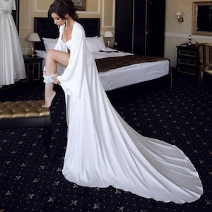 May include: A bride in a white satin robe with a long train, standing in a hotel room, putting on a garter.