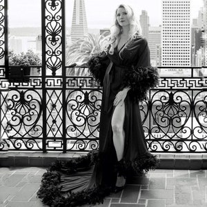 May include: A woman wearing a black robe with feather trim. The robe is open in the front and has a long train. The woman is standing on a balcony with a city view in the background.