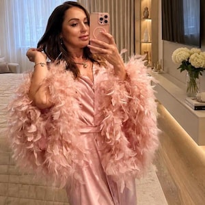 May include: A light pink satin robe featuring long sleeves adorned with voluminous matching pink feathers. The garment has a V-neckline, a wrap-style front with a tie waist, and a smooth, lustrous finish that reflects light across the fabric surface.