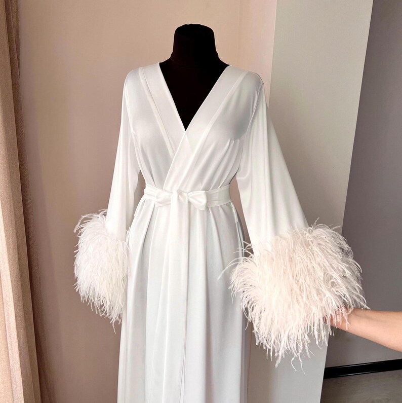 May include: A white satin robe with long sleeves and a V-neckline. The sleeves have white feather trim.