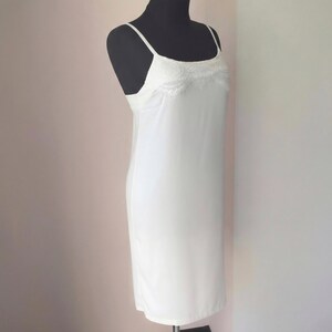 May include: A white slip dress with spaghetti straps and a lace trim detail at the neckline. The dress is displayed on a black mannequin. The slip dress is a classic piece of lingerie.
